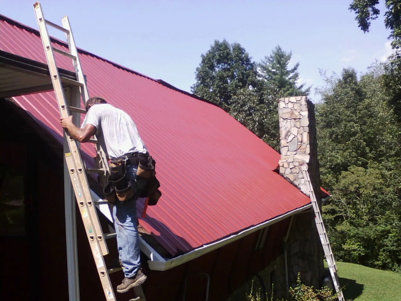 Roofer on a ladder beside a red metal roof and stone chimney — Soffit & Fascia Repair by Roofing Brothers in Four Oaks